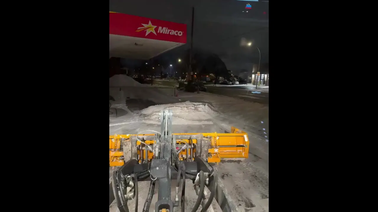 Pousseur à neige repoussant la neige dans un stationnement commercial la nuit, sous l'éclairage d'une station-service